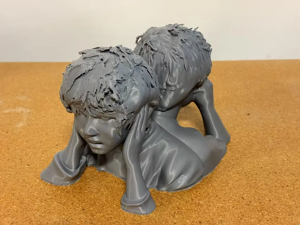 灰色3D打印双人半身像，头靠双手，置于软木表面，从左前方拍摄。