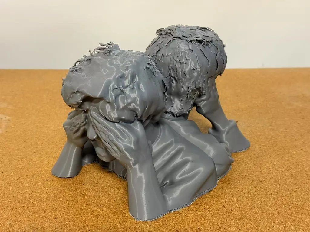 灰色3D打印双人半身像置于软木表面，近处头部遮脸，从左侧拍摄。