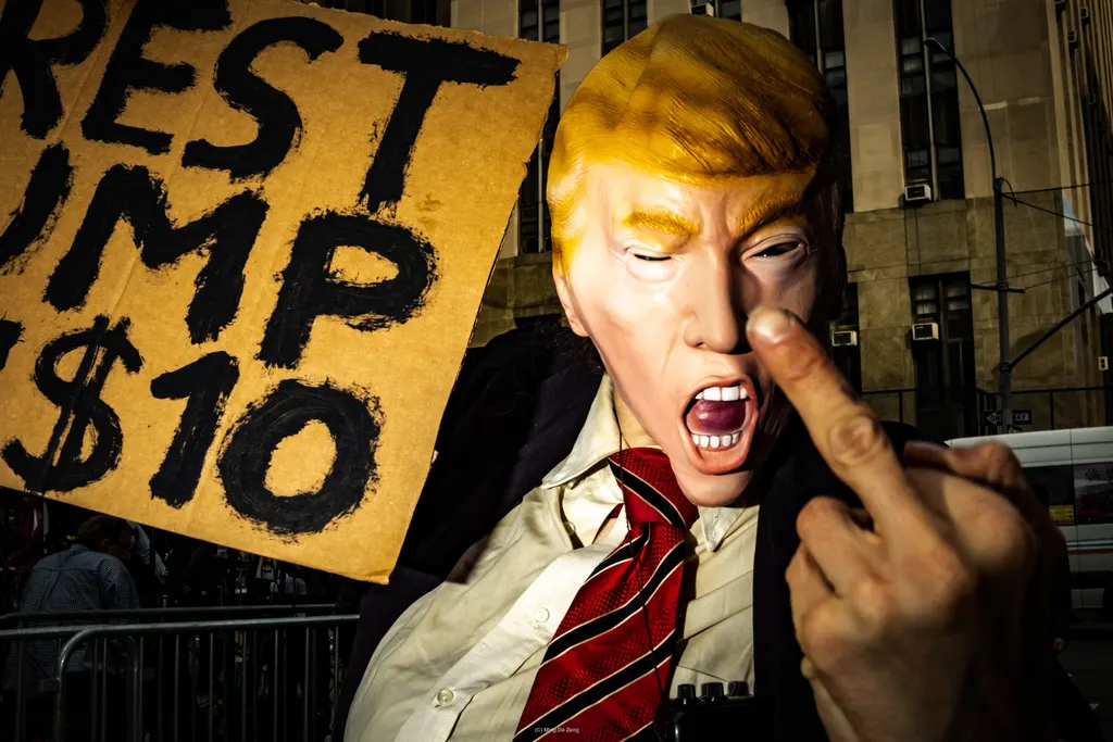 特朗普面具朝镜头伸手，旁边纸板牌写着“Arrest Trump $10”。