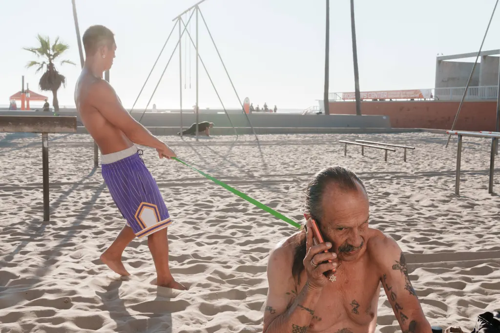 海滩健身区内，前景男子打电话，后方另一名男子拉着绿色弹力带。