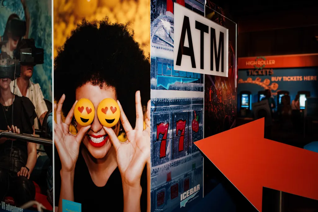 室内游乐场标牌写着“ATM”，有老虎机图案、VR乘客和一个大型橙色箭头。