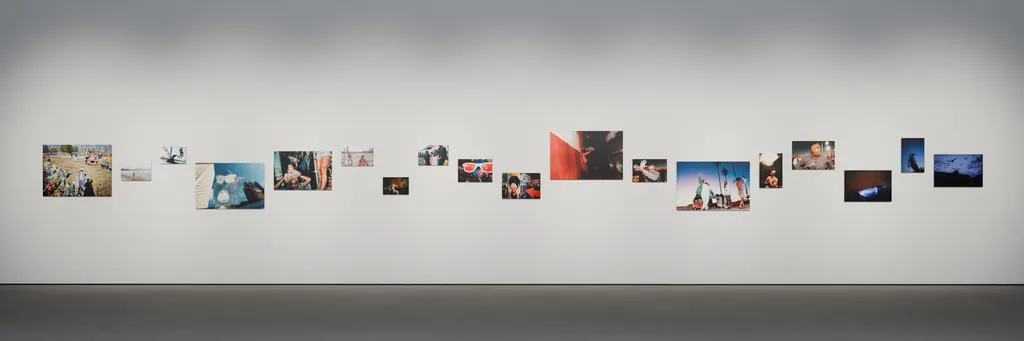 Installation rendering with eighteen photographs arranged at varying heights as a continuous frieze across a gallery wall.