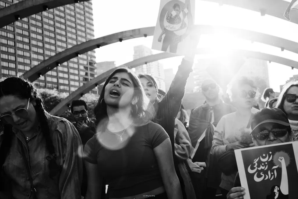 Black-and-white marchers under arched beams; a woman chants while others hold signs, including one with Persian text.