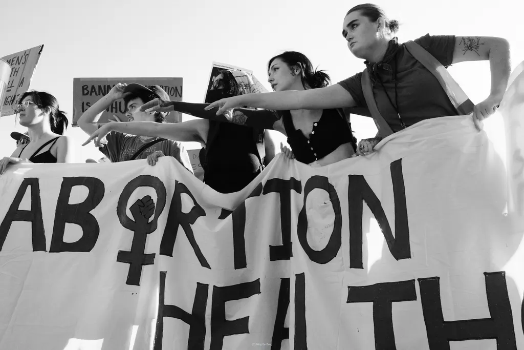 Black-and-white protesters hold a large banner with “ABORTION” visible; several people point forward from behind it.