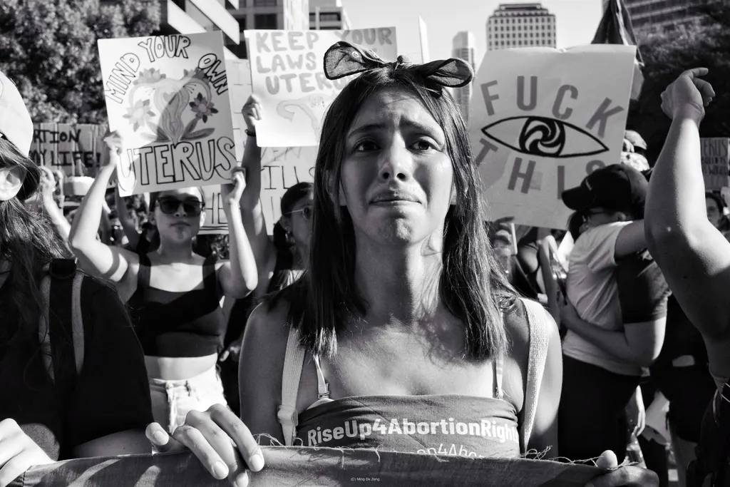 Black-and-white protest crowd; a young woman holds a banner as signs behind read about uterus laws and “FUCK THIS.”