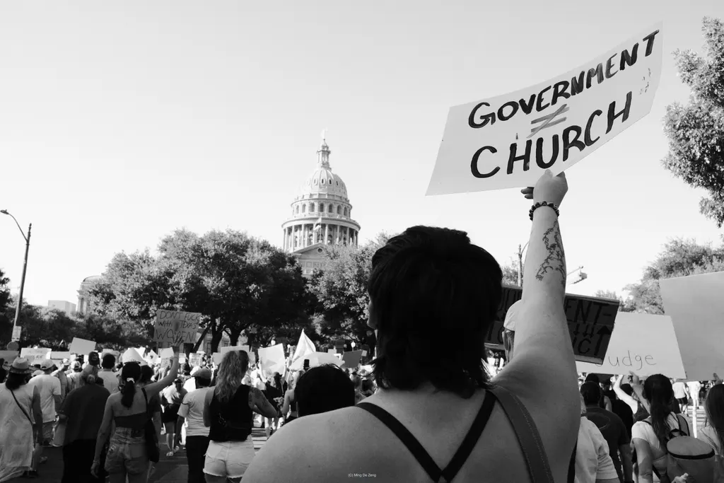 Black-and-white crowd near a capitol dome; a person seen from behind raises a sign reading “Government ≠ Church.”