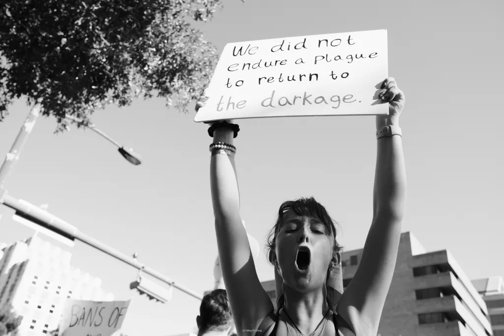 Black-and-white photo of a woman shouting with both arms raised, holding a sign about not returning to “the dark age.”