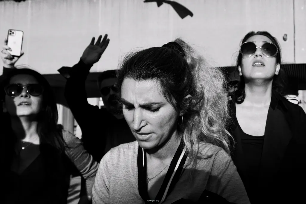 Black-and-white close crowd scene; a central woman looks down while others wear sunglasses, one holding up a phone.
