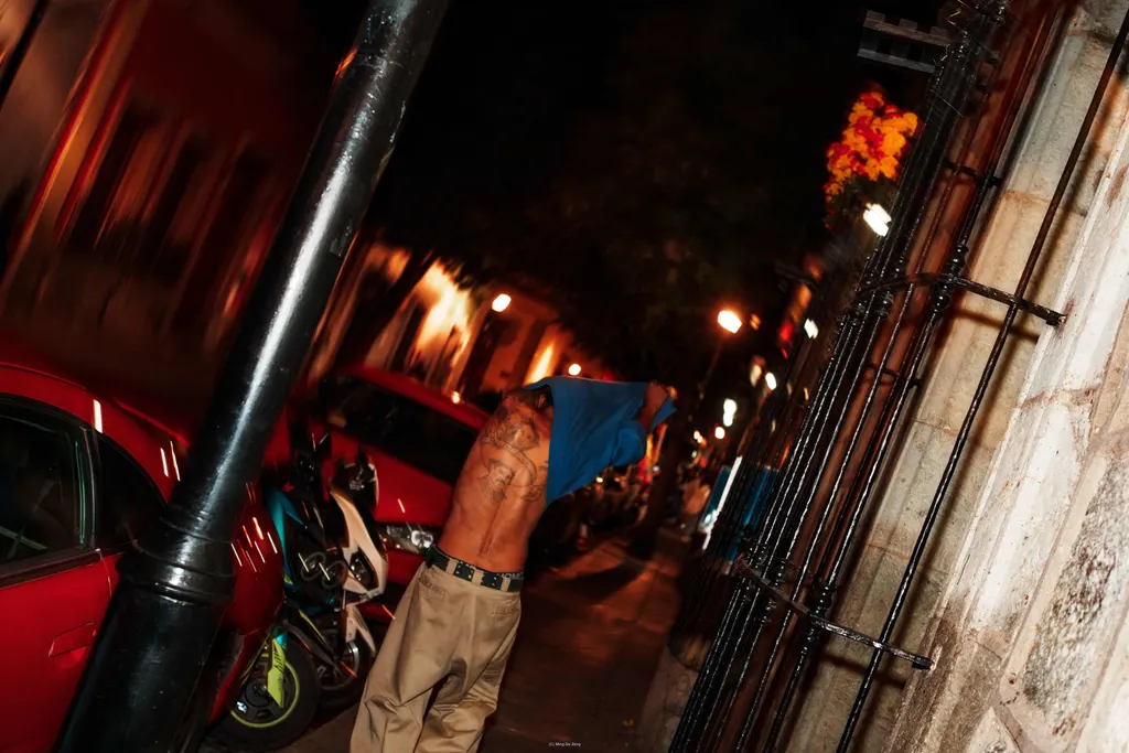 倾斜的夜间街景中，一名赤裸上身男子在停放车辆旁把蓝色上衣套过头。