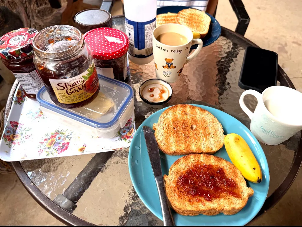 玻璃桌上的早餐，有吐司、果酱罐、香蕉、杯子、餐刀和黑色手机。