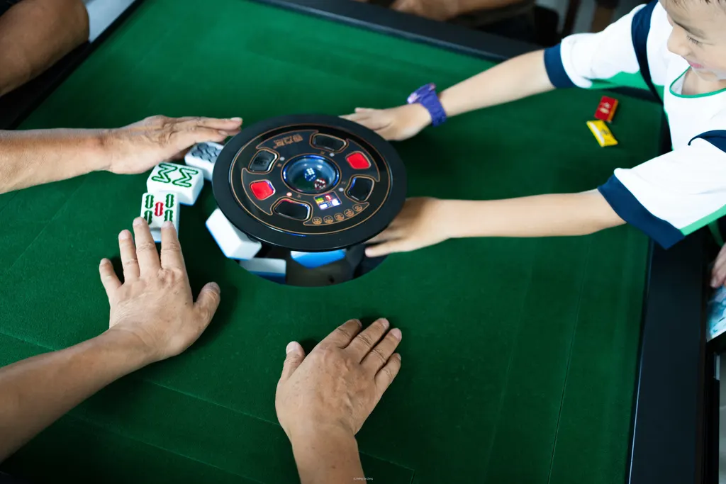Hands reach around a green mahjong table with an automatic tile machine in the center.
