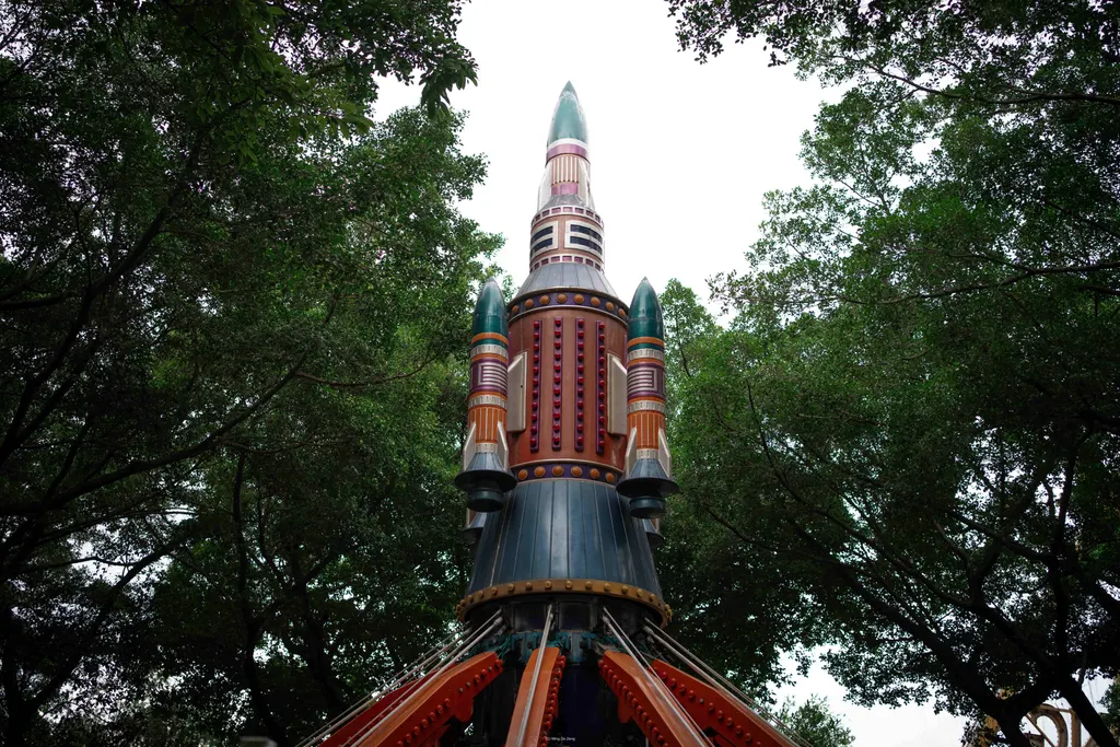 Tall rocket-shaped amusement ride rises among dense green trees.