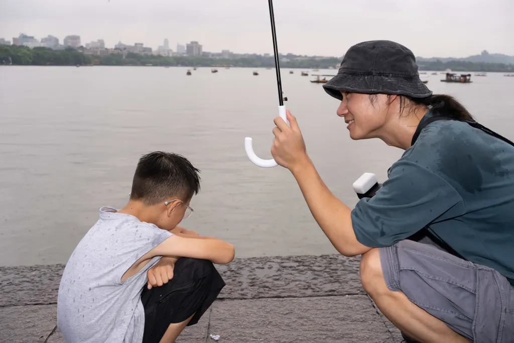 Adult crouches with an umbrella beside a seated boy on a lakeside walkway.