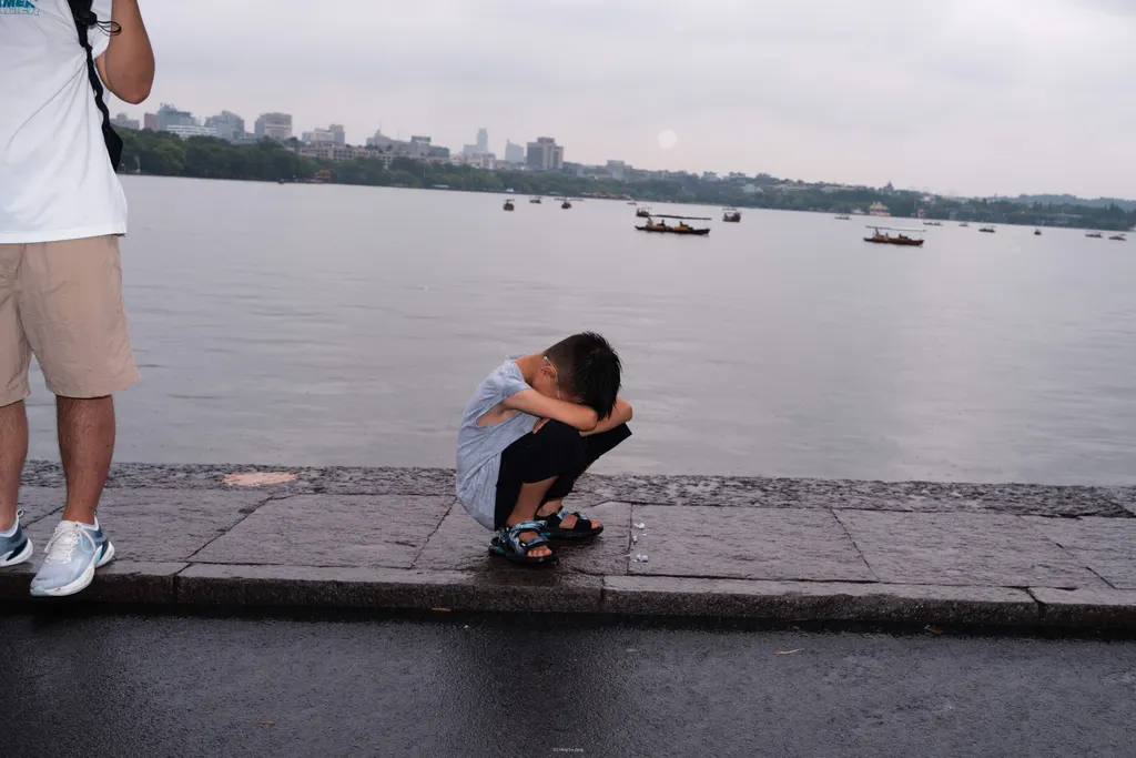 Boy crouches by a lakeside curb, with an adult's legs at left and boats in the distance.