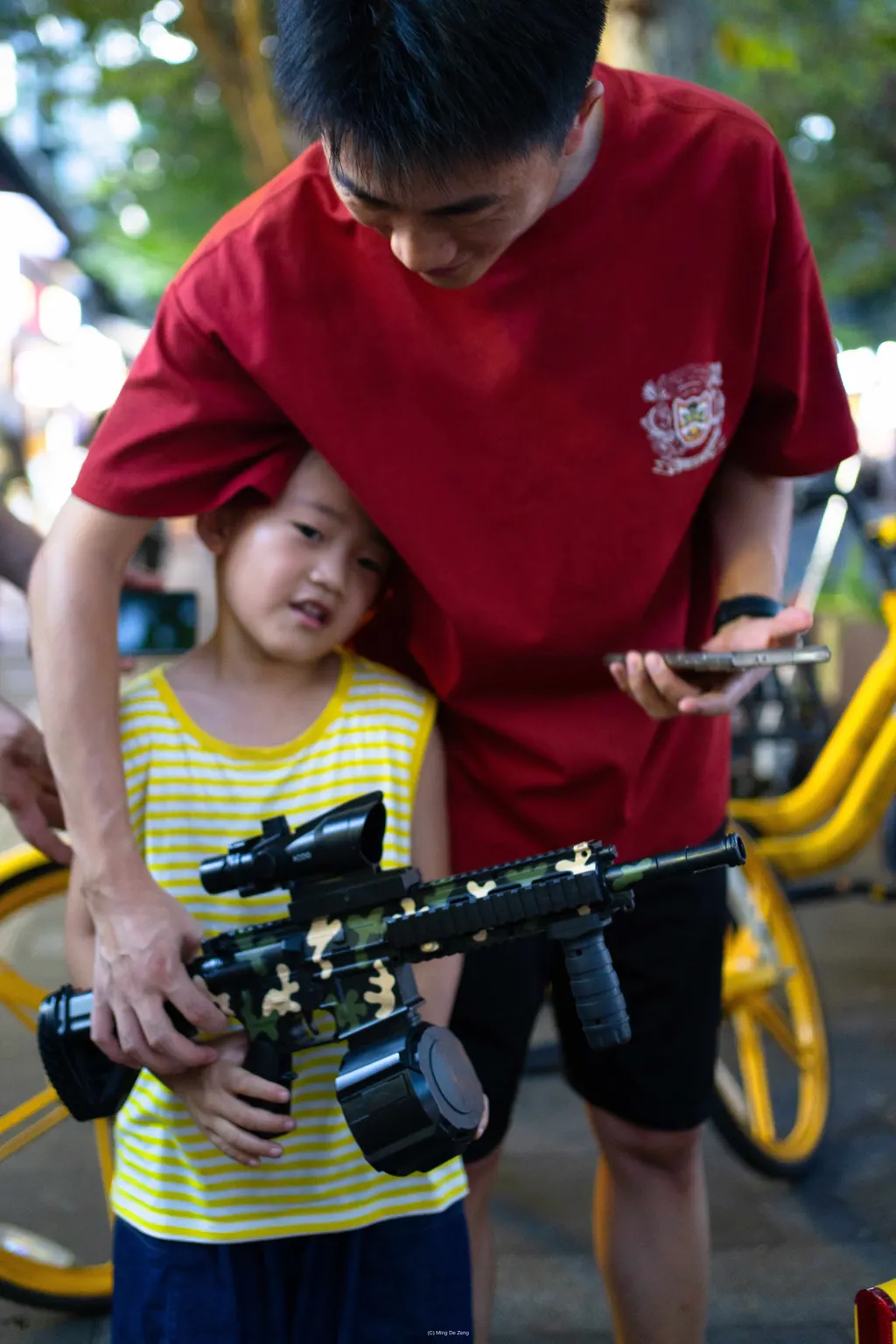 Child holds a toy rifle while an older boy in a red shirt stands close with a phone.