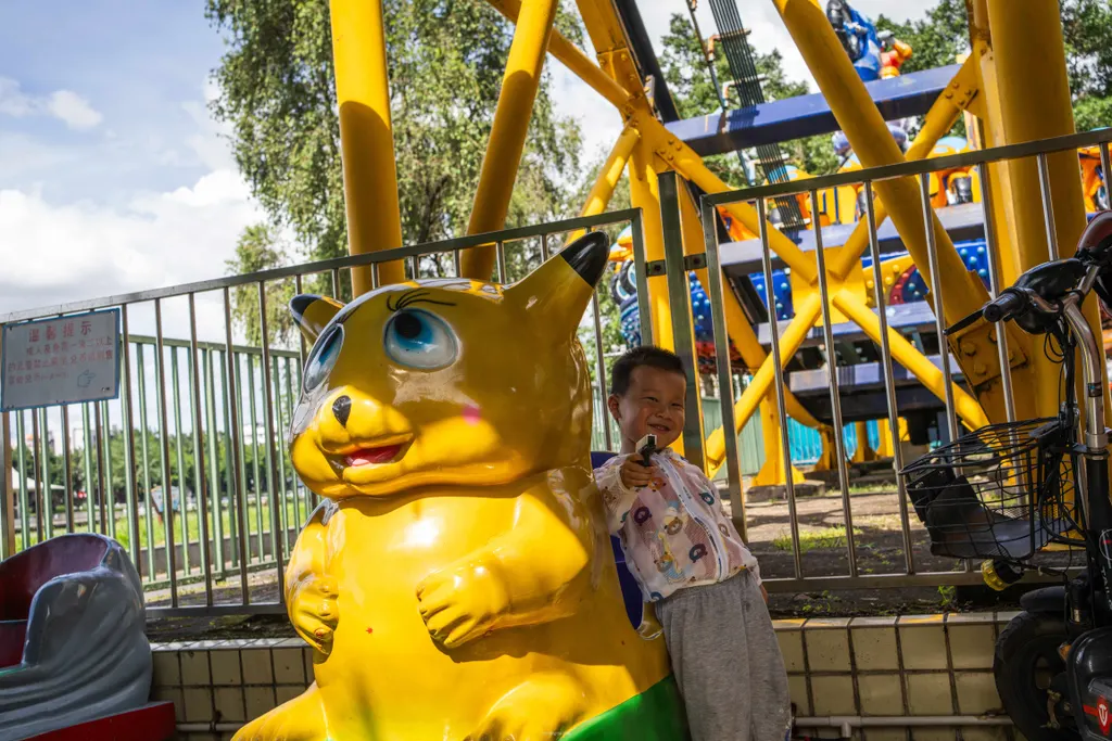 Boy smiles beside a yellow animal ride figure, pointing a small toy gun toward the camera.