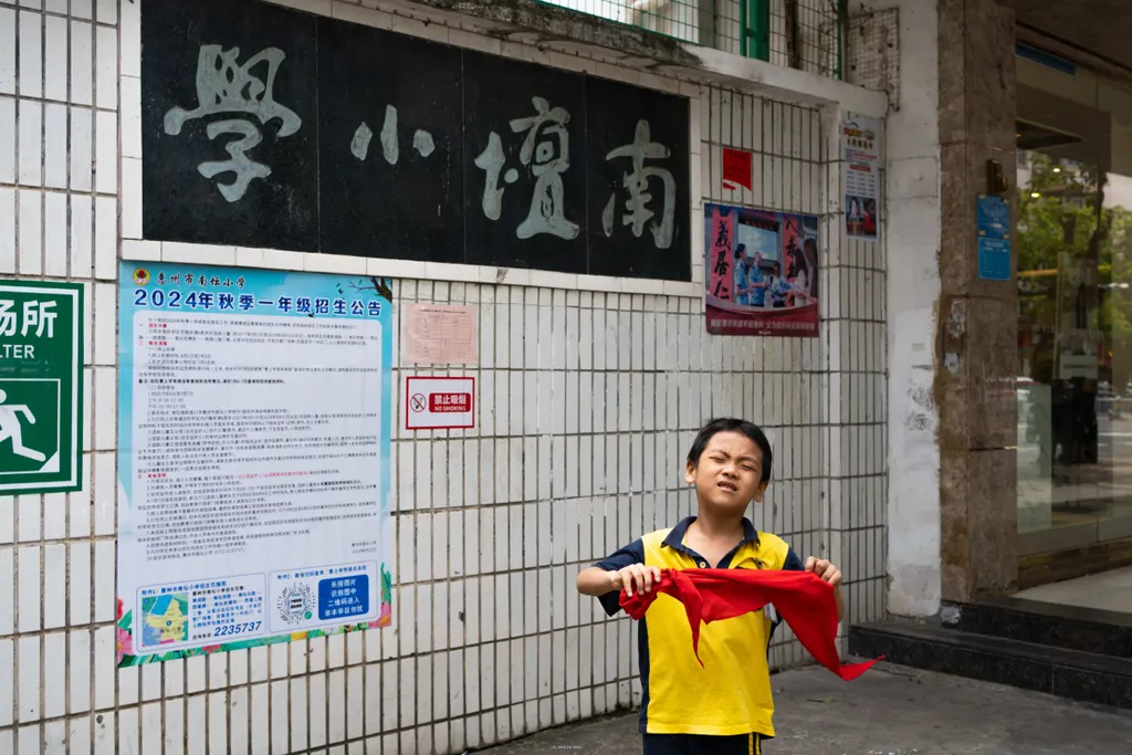 Boy holds red cloth below a Chinese school sign and a 2024 enrollment poster.