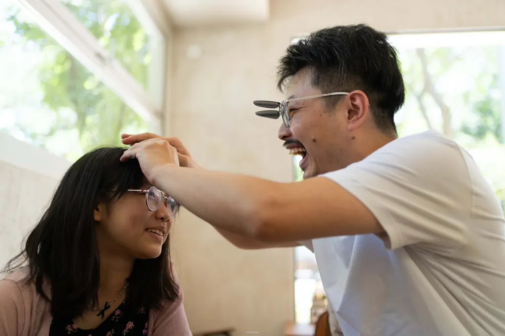 Man in glasses laughs while leaning toward a girl indoors, hands near her head.