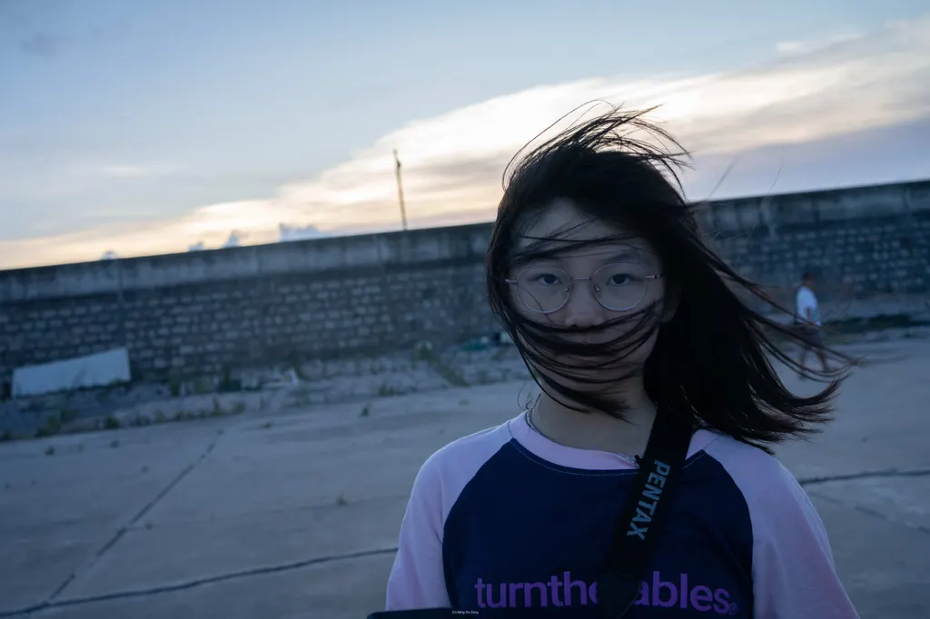 Girl with glasses stands outdoors at dusk, wind blowing hair across her face.