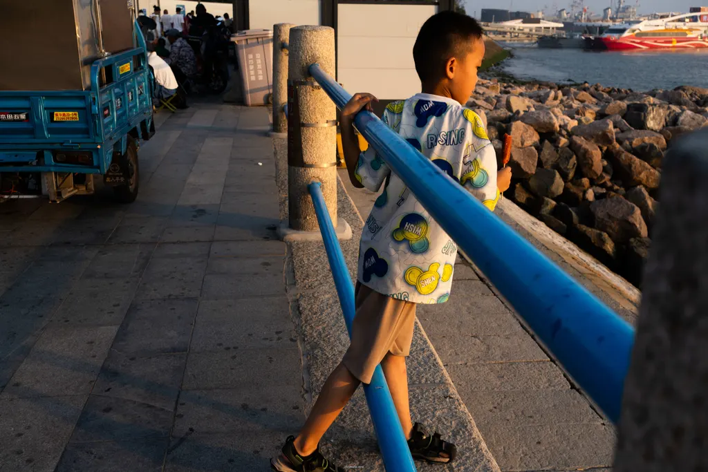 Boy leans on a blue rail eating a snack beside rocks and water at sunset.