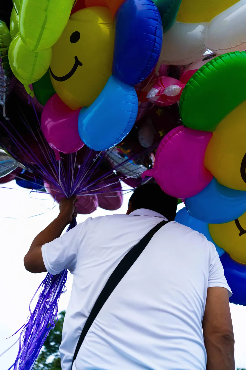 Balloon seller seen from behind carrying clusters of colorful balloons with smiley faces.
