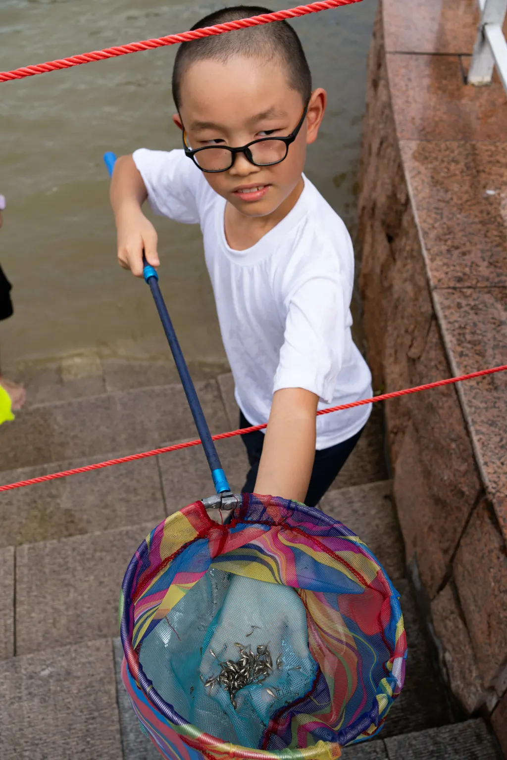 Boy with glasses holds a fishing net over steps by the water, framed by red ropes.