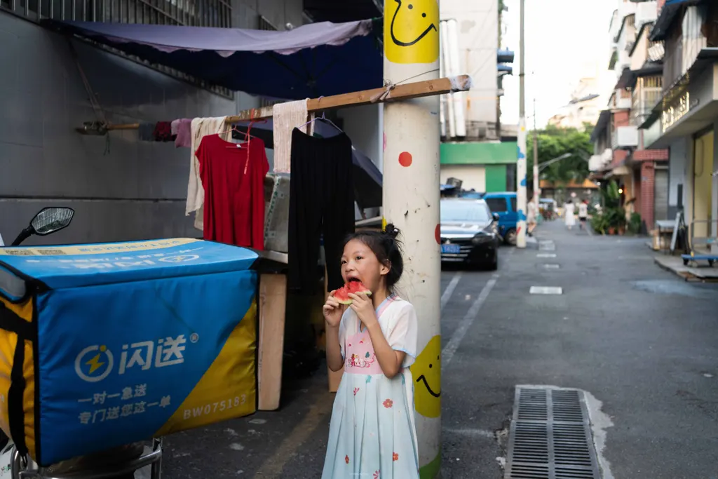 Girl eats watermelon on a street beside a blue delivery scooter and a pole with smiley stickers.