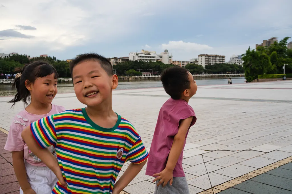 Three children stand on a tiled plaza, one smiling toward the camera in a striped shirt.